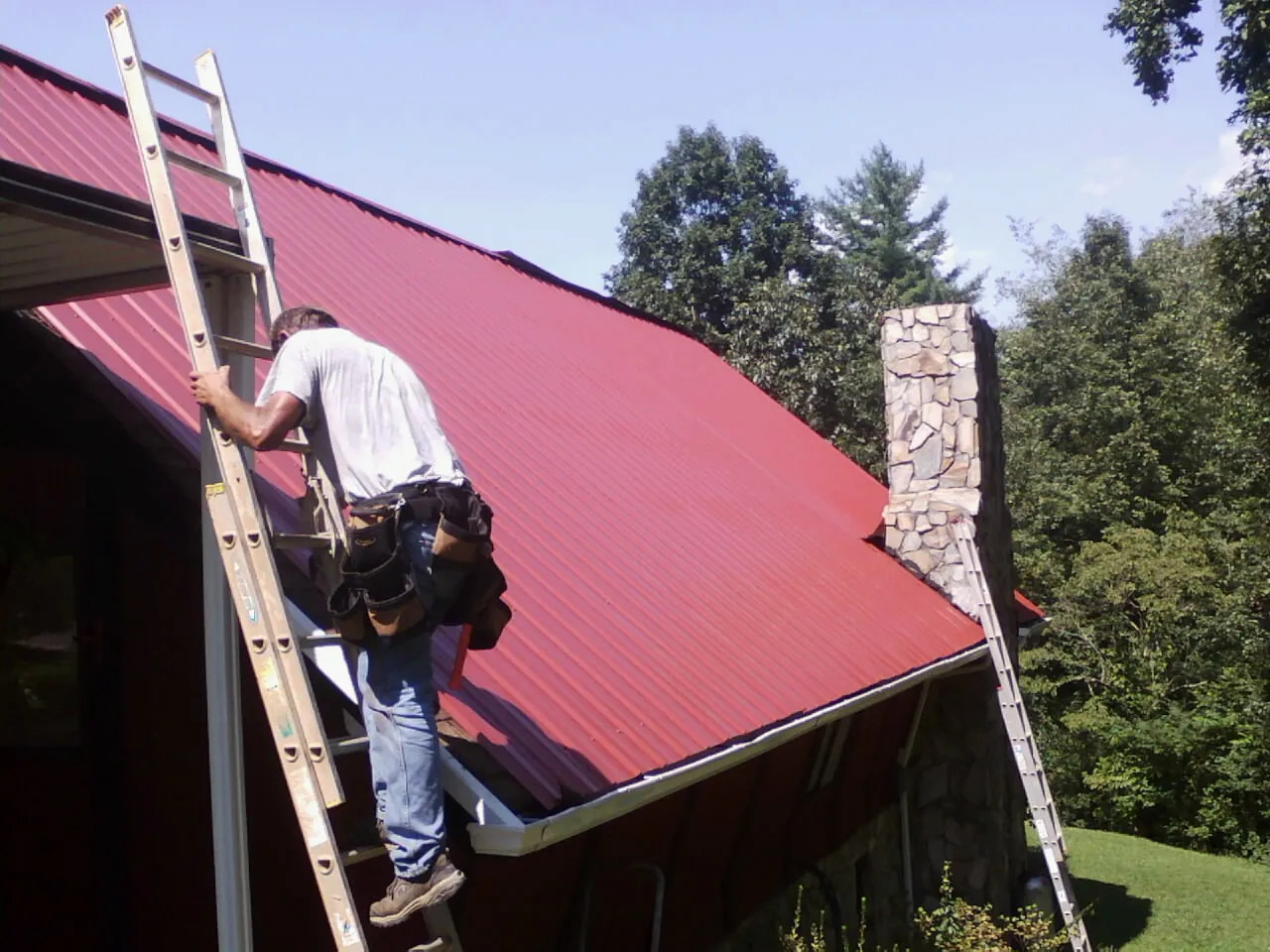 Roofer on a ladder beside a red metal roof and stone chimney — Soffit & Fascia Repair by Roofing Brothers in Bay Minette